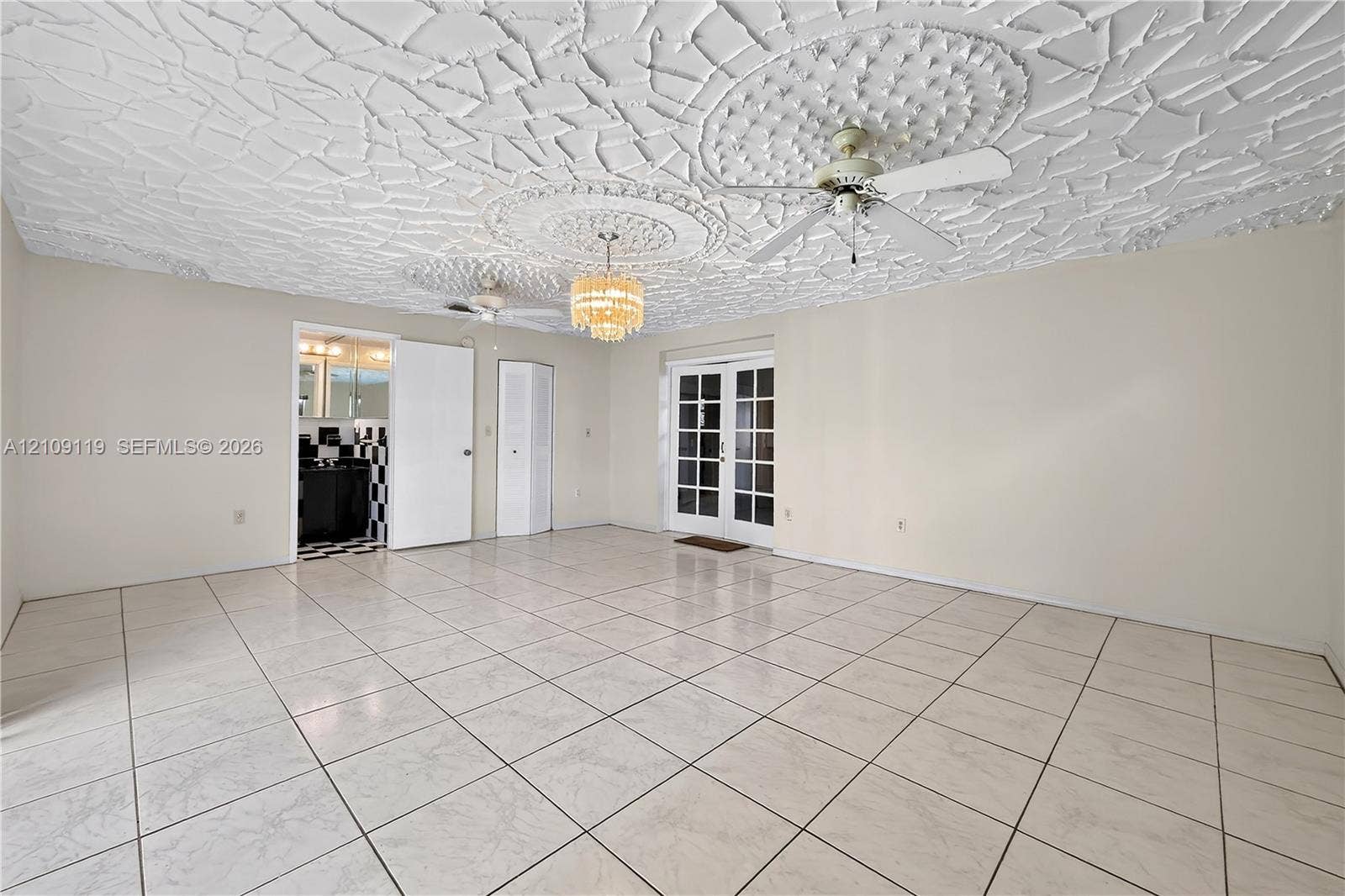 Spacious room featuring a decorative plaster ceiling with medallion accents, large-format tile flooring, a tiered crystal chandelier, a white ceiling fan, and French doors