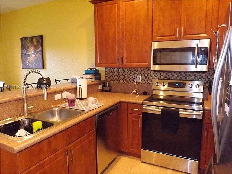 2529 SW Stockbridge Square SW, Vero Beach, FL 32962 Kitchen featuring stained wood cabinetry, stainless steel appliances, a multi-tone tile backsplash, and a double-basin sink with a gooseneck faucet