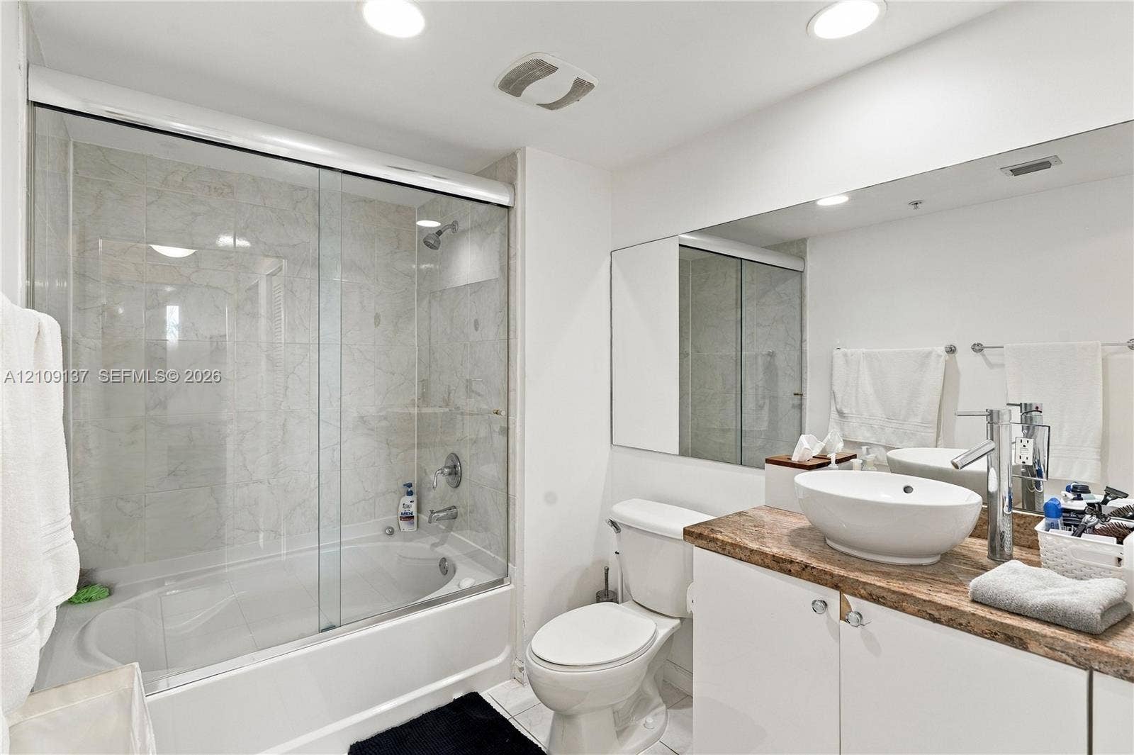 18800 NE 29th Avenue, #411, Aventura, FL 33180 Bathroom featuring a vessel sink atop a stone-finish countertop, white cabinetry, a full-width mirror, and a soaking tub with a sliding glass enclosure and tiled surround