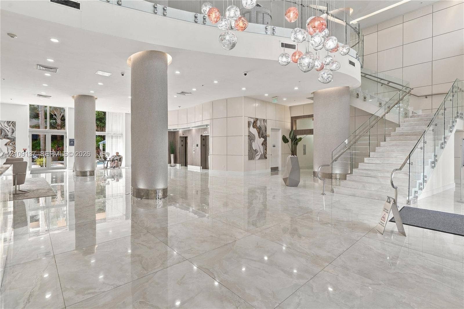 18800 NE 29th Avenue, #411, Aventura, FL 33180 Grand lobby featuring polished stone tile flooring, structural columns with metallic-finish cladding, a multi-globe pendant chandelier, and a glass-paneled staircase