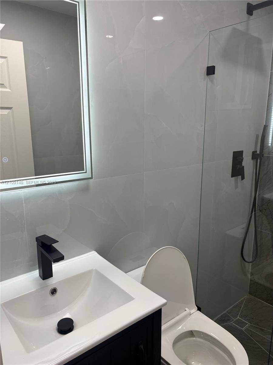 Floating vanity with a white countertop, wall-mounted illuminated mirror, frameless glass shower enclosure, and matte black fixtures