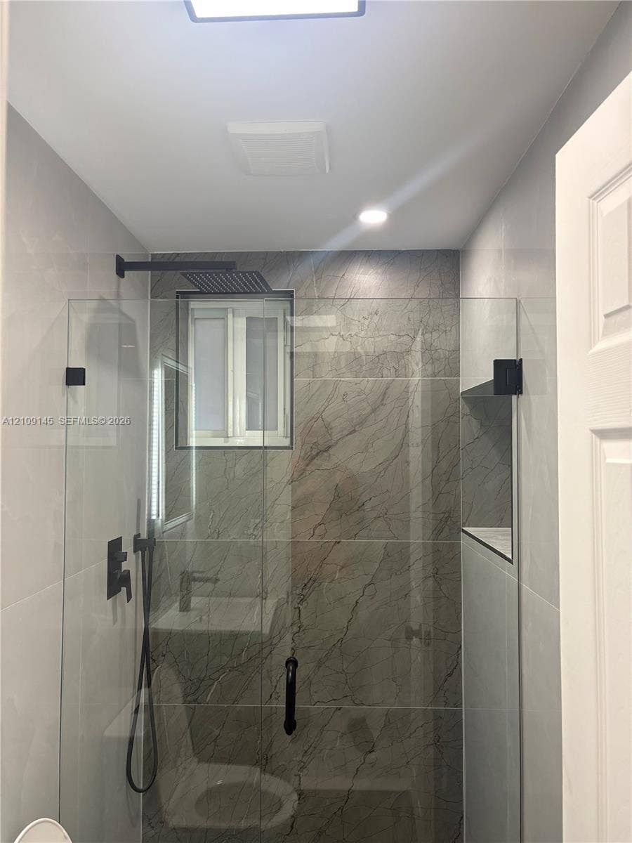 Walk-in shower featuring large-format stone-pattern tile surround, frameless glass enclosure, matte black rainfall shower head, handheld sprayer, and built-in recessed shelf