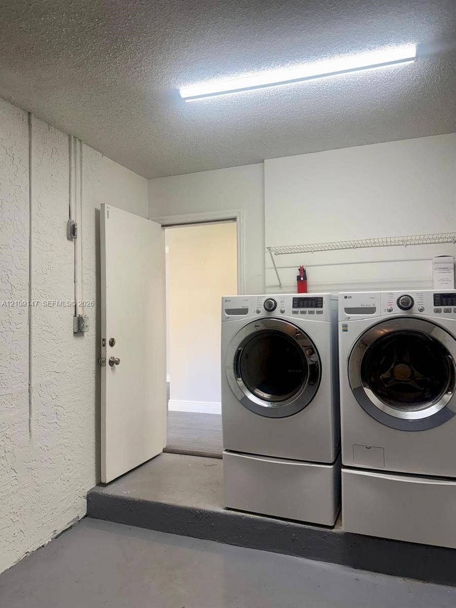 Laundry area featuring white front-load washer and dryer on pedestals, overhead wire shelving, textured wall finish, and painted concrete flooring