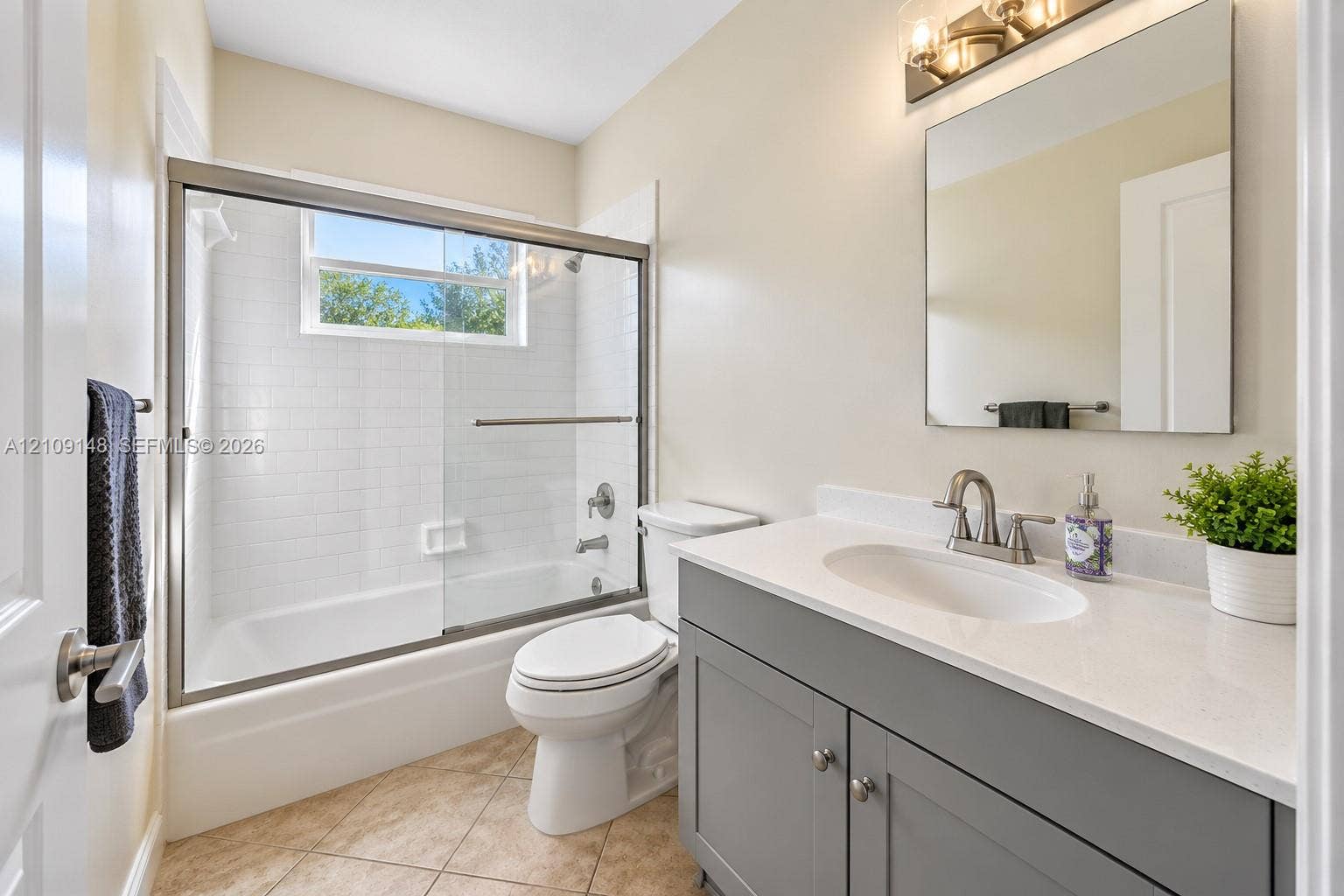 1701 SW 23rd Avenue, Fort Lauderdale, FL 33312 Bathroom featuring a gray vanity with a neutral solid-surface countertop, an undermount oval sink, a frameless mirror with mounted lighting, and a bathtub with a white subway-tile surround and a sliding glass enclosure