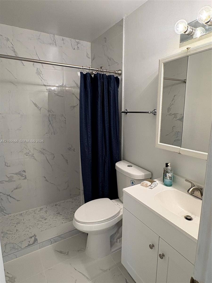 Bathroom featuring shower walls and flooring finished in white stone-pattern tile, white vanity cabinet with integrated basin, polished metal faucet, and wall-mounted medicine cabinet with integrated lighting