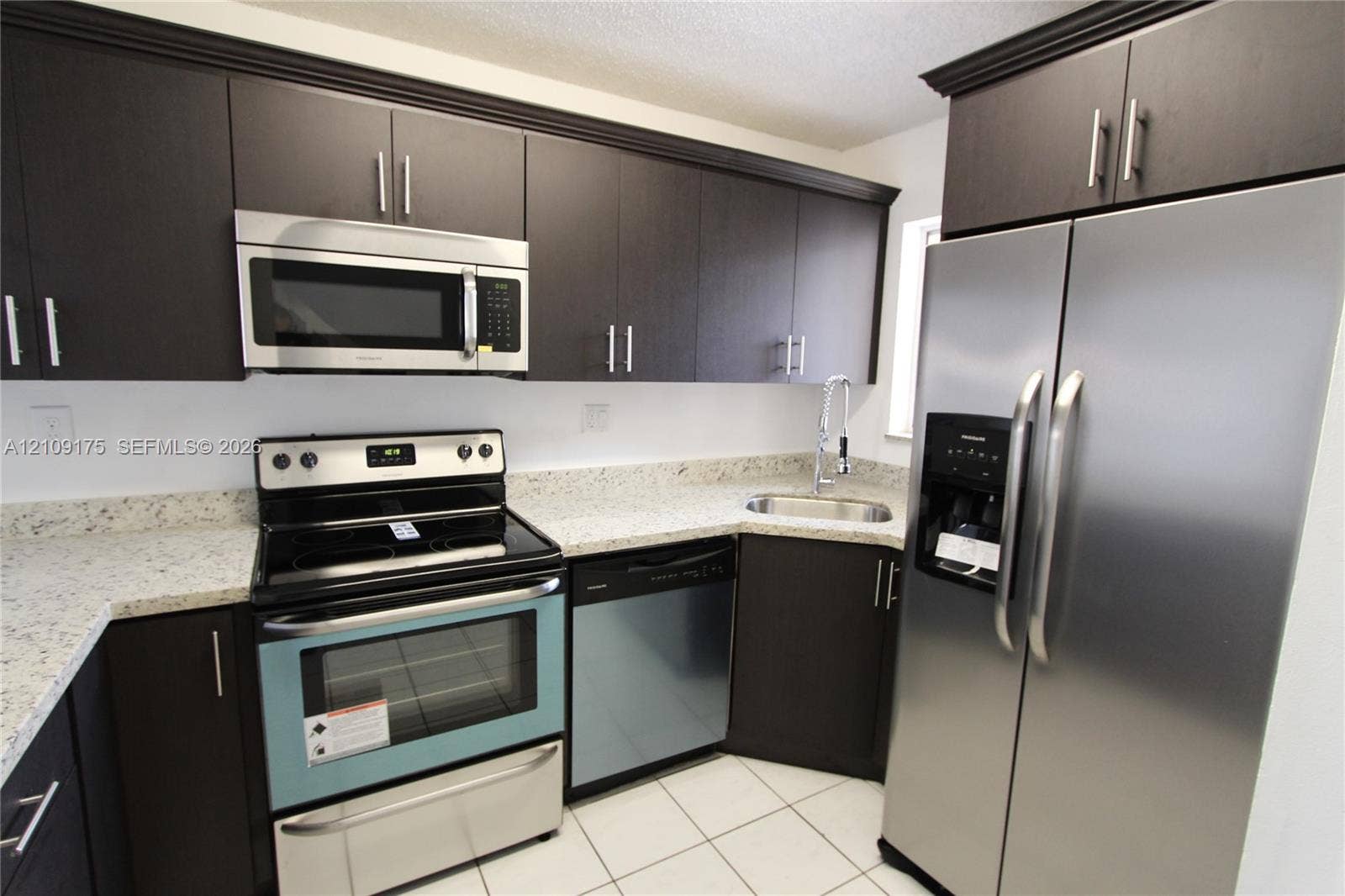 Dark-toned cabinetry with metal hardware, light-toned stone-pattern countertops, stainless steel appliances, and tile flooring