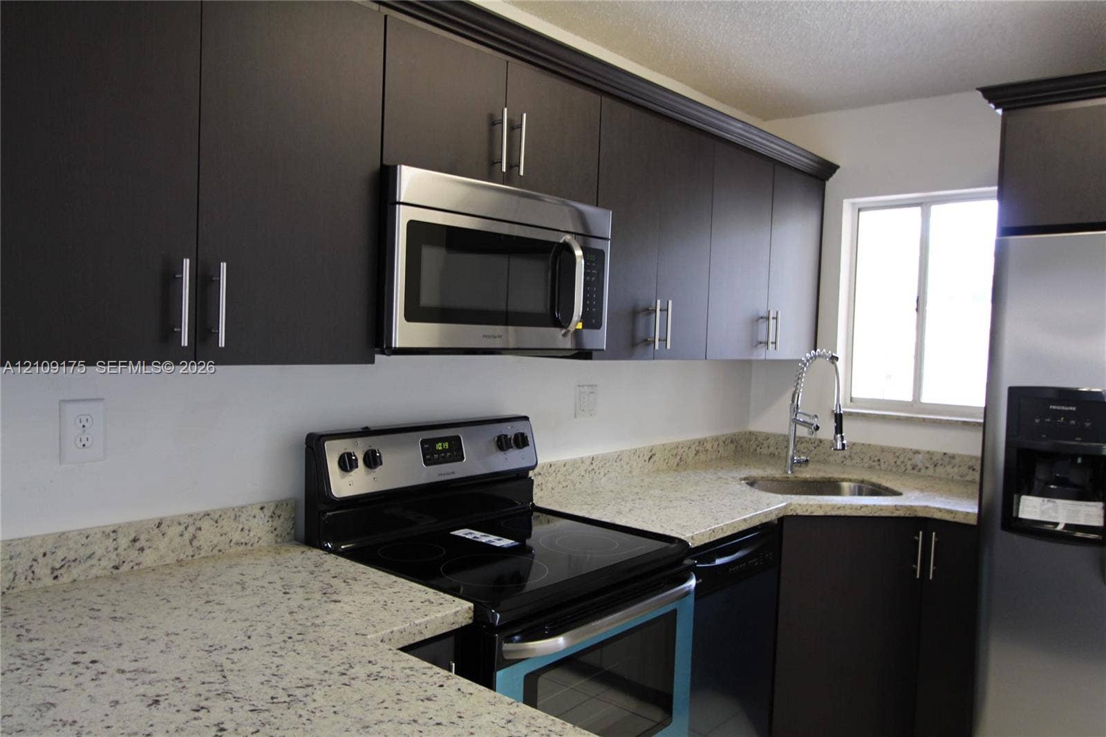Kitchen featuring dark-toned cabinetry, light stone-patterned countertops, stainless steel appliances, a corner-mounted sink with a pull-down faucet, and crown molding