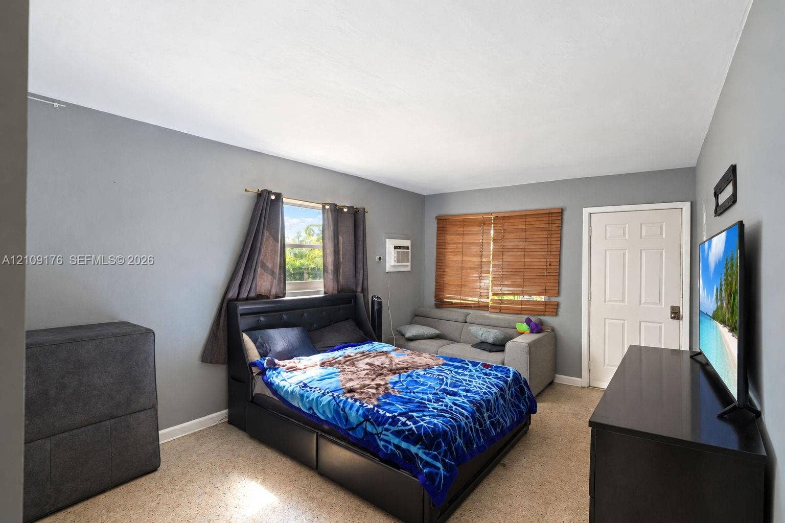 Interior space featuring neutral grey wall paint, textured floor finishes, dual windows with wood-slat blinds, and a wall-mounted climate control unit