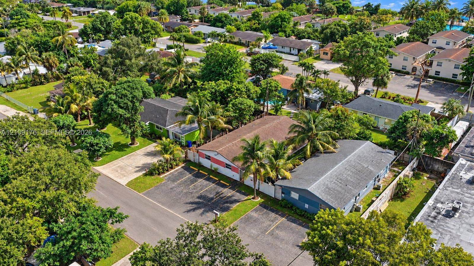 Aerial perspective of the property featuring a paved off-street parking area, multi-tone exterior siding, and a shingled roof, surrounded by mature palm trees and established landscaping