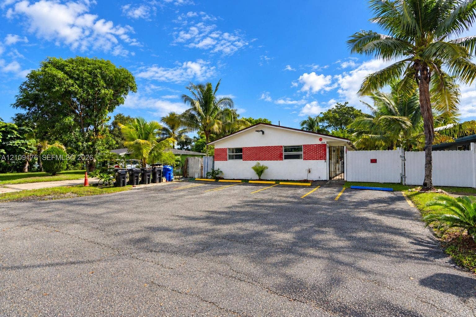 Single-story exterior featuring white stucco walls with red masonry accents, paved off-street parking area, and white vinyl privacy fencing