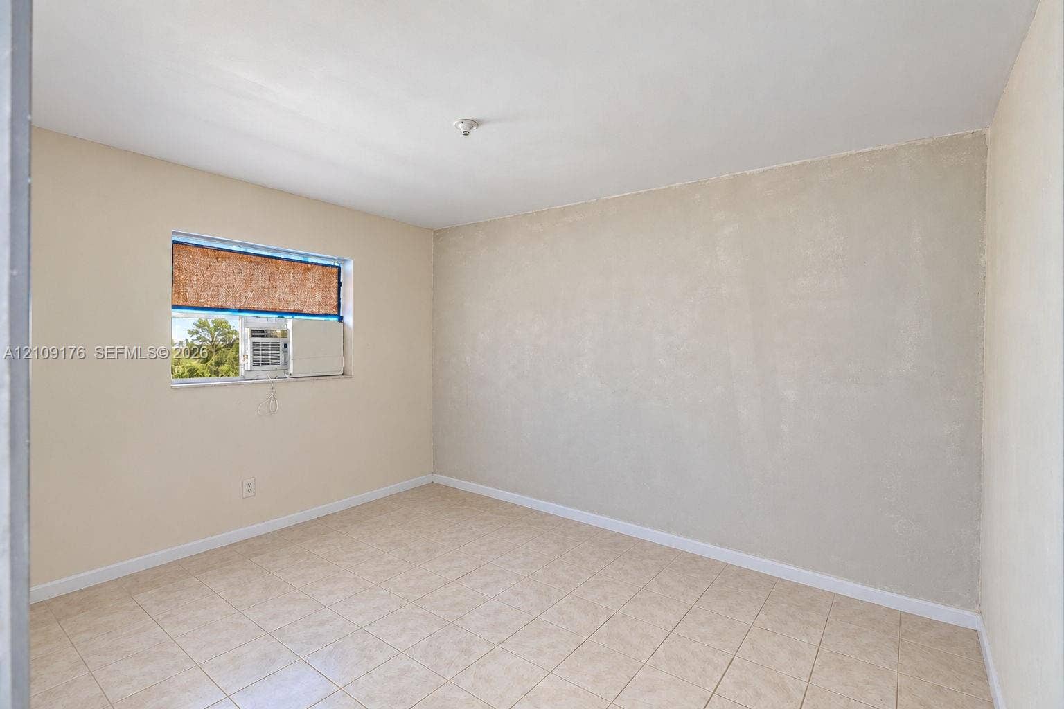 Neutral-toned interior room featuring tile flooring, smooth walls, and a single window with a built-in air conditioning unit