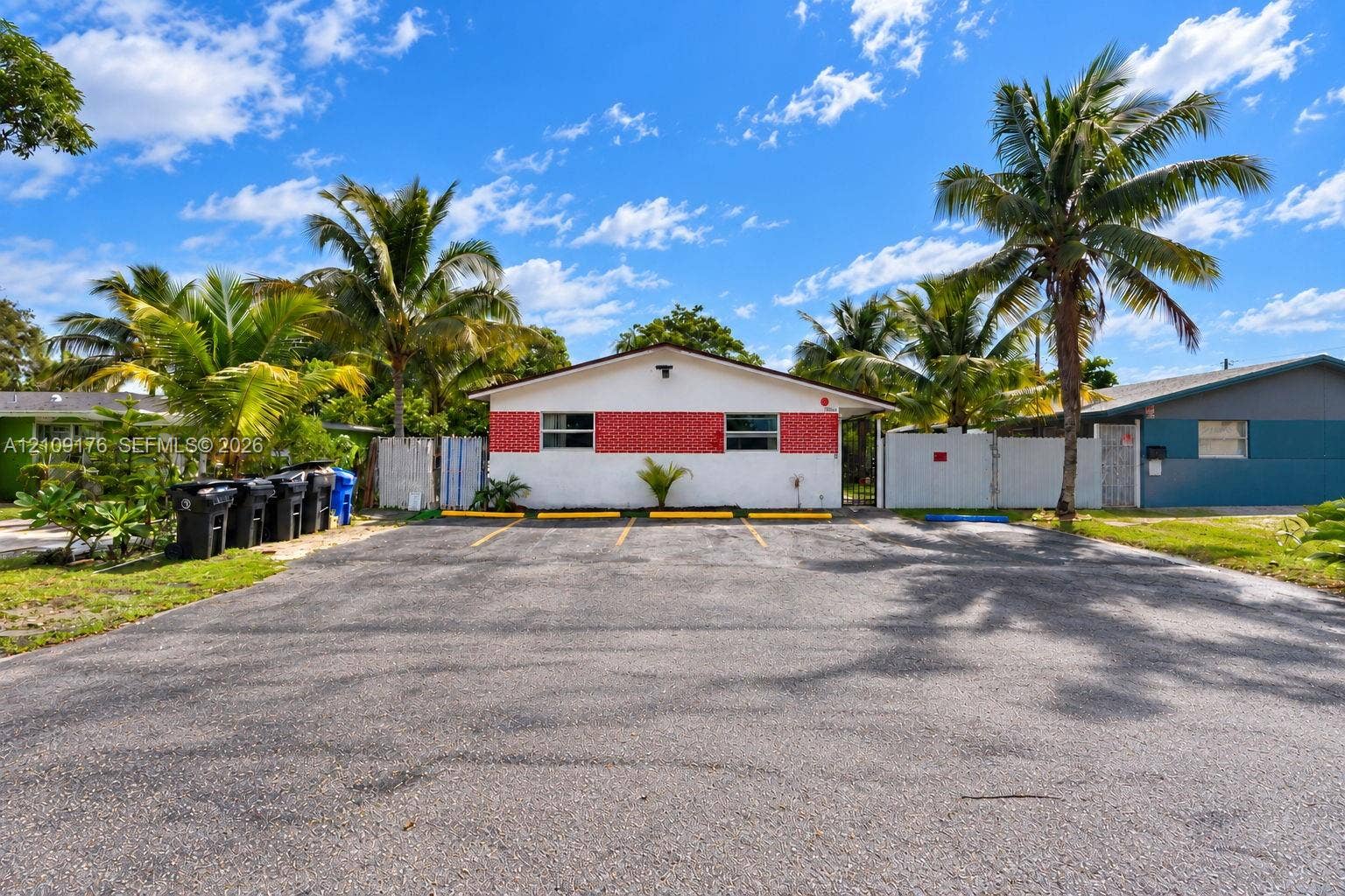 Exterior featuring a white facade with red masonry accents, a paved parking area with designated spaces, and mature palm trees