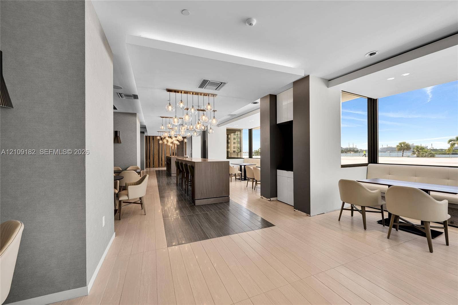 Open-concept interior featuring light-toned tile flooring, dark wood-finish accent flooring, decorative multi-pendant lighting, and expansive windows