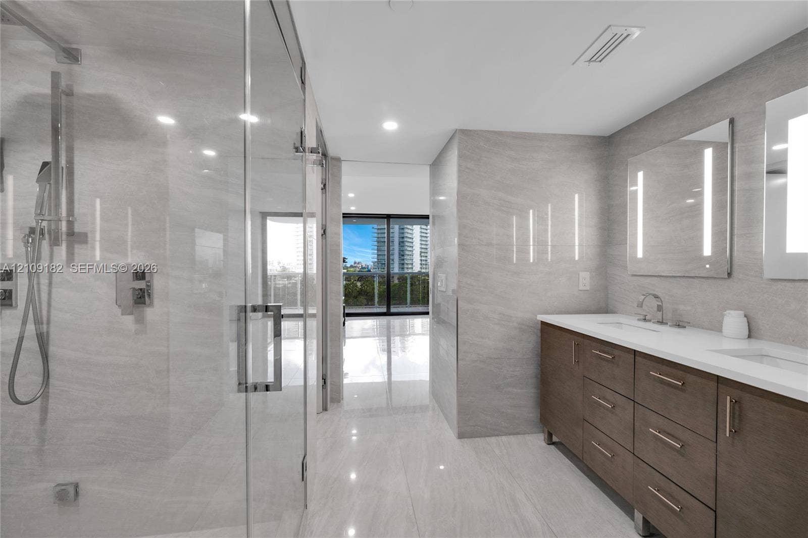 Double vanity with wood-finish cabinetry, white countertop, integrated dual sinks, and frameless glass-enclosed shower with tiled wall surfaces