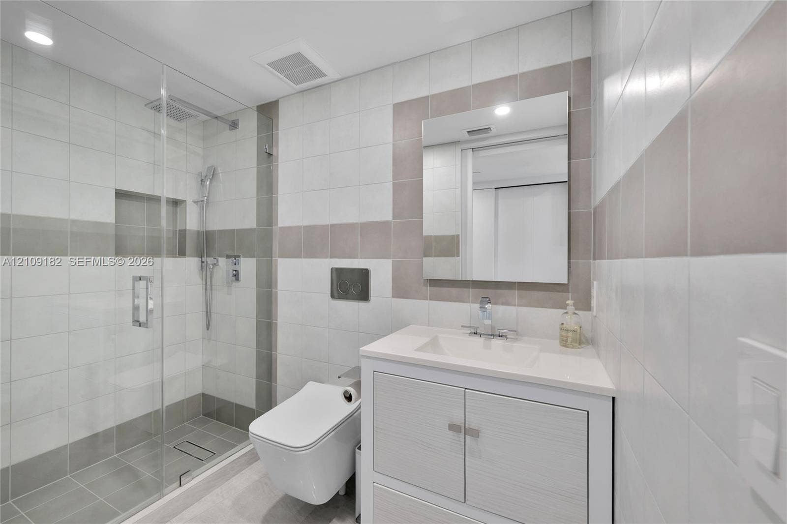 Bathroom featuring a walk-in glass-enclosed shower, wall-mounted dual-flush toilet, vanity with integrated basin, polished chrome fixtures, and neutral-toned wall tile