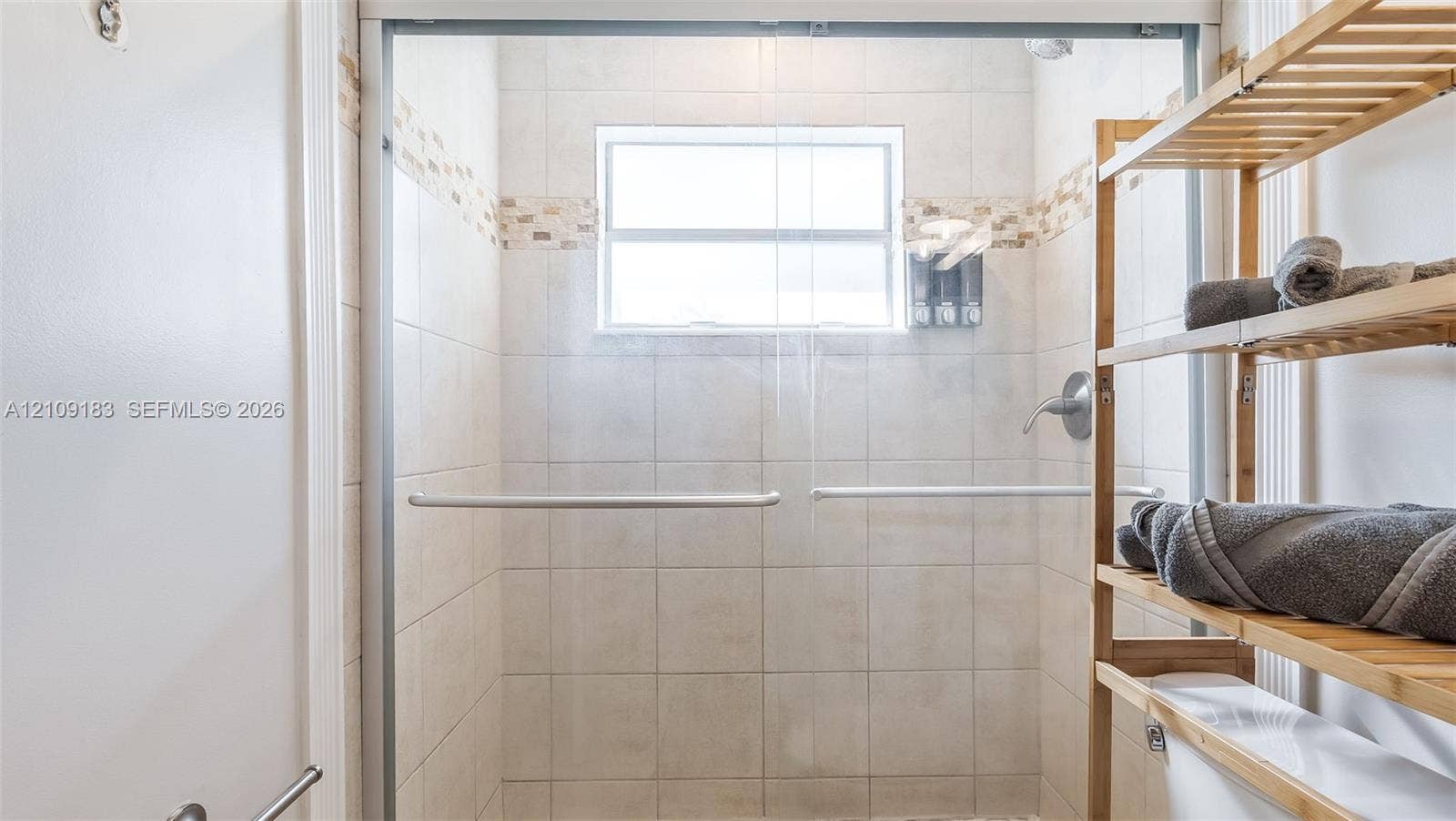 1009 NE 17th Court, Fort Lauderdale, FL 33305 Walk-in shower enclosure featuring light-toned ceramic wall tile, horizontal mosaic tile accent banding, a framed glass door, and a built-in window