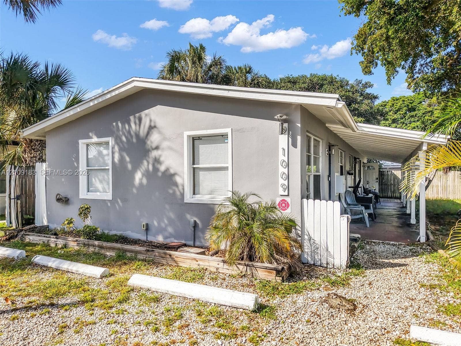 1009 NE 17th Court, Fort Lauderdale, FL 33305 Exterior featuring a light grey stucco finish, a covered concrete patio, white picket fencing, and a gravel parking area with concrete wheel stops