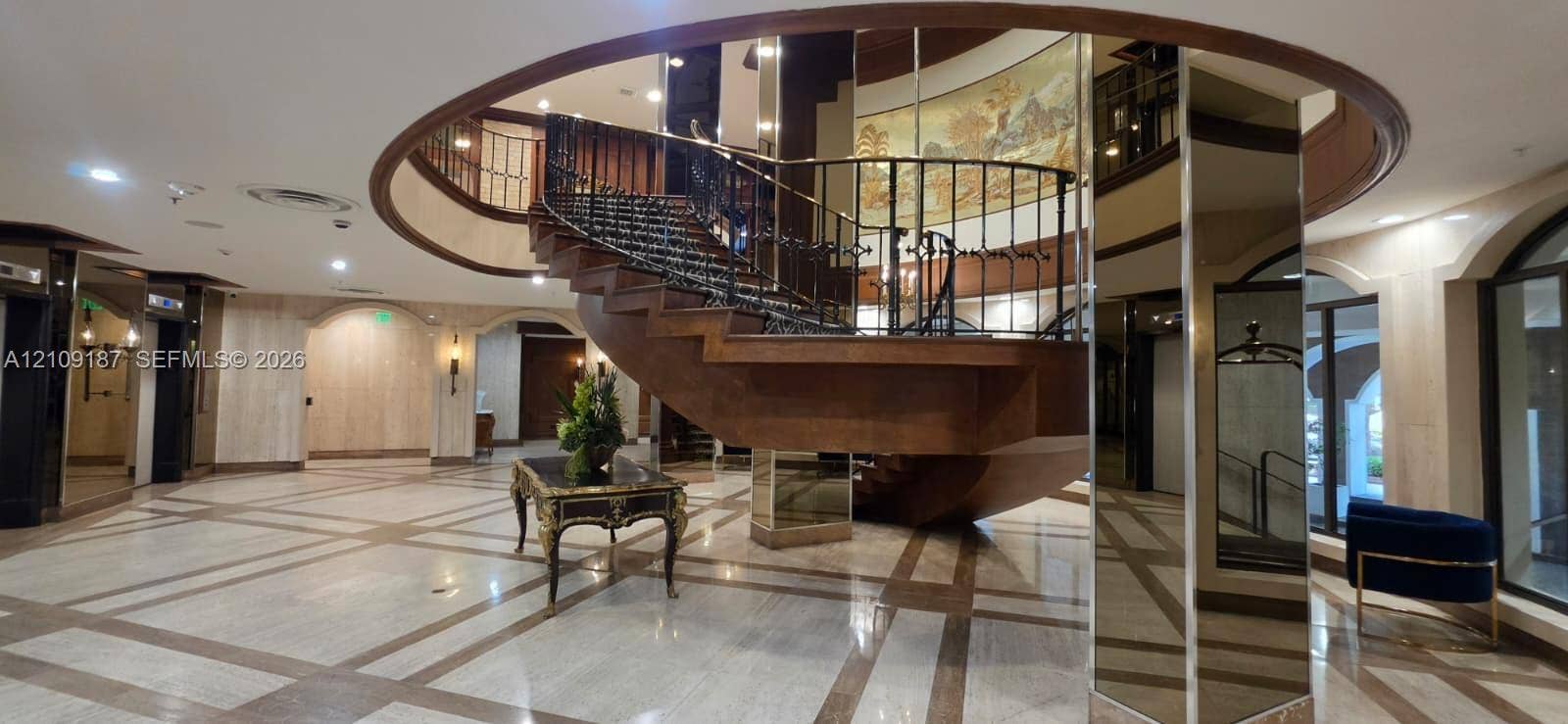 6039 Collins Avenue, #1625, Miami Beach, FL 33140 Grand foyer featuring a circular architectural cutout, a sweeping wooden staircase with a metal balustrade, patterned stone flooring, and mirrored support columns