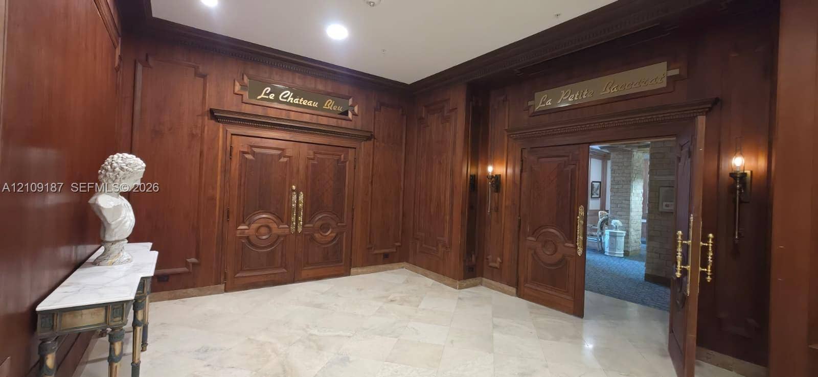 6039 Collins Avenue, #1625, Miami Beach, FL 33140 Elegant great room Featuring wood-paneled walls, intricate double wooden doors, marble flooring, and wall-mounted brass light fixtures.
