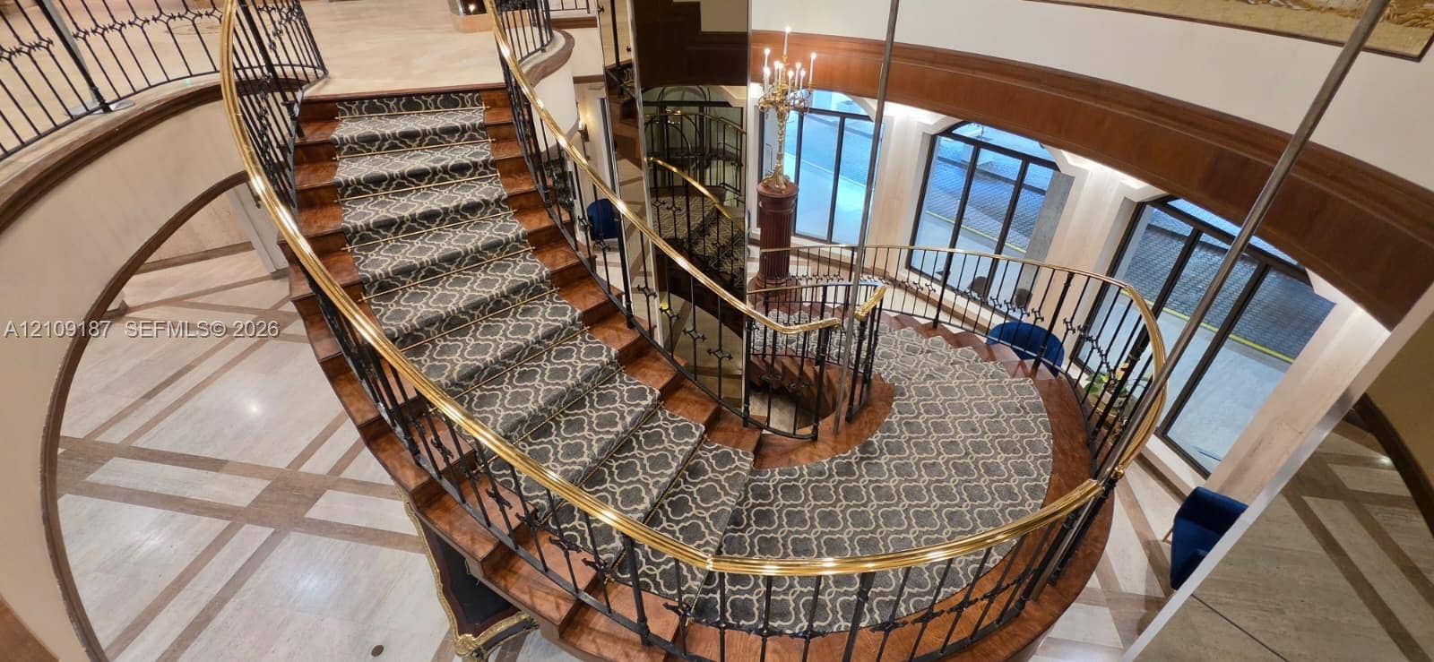 6039 Collins Avenue, #1625, Miami Beach, FL 33140 Curved staircase featuring wood-finish treads, custom patterned carpet inlay, wrought iron balusters, and a brass handrail, complemented by stone tile flooring with decorative inlay