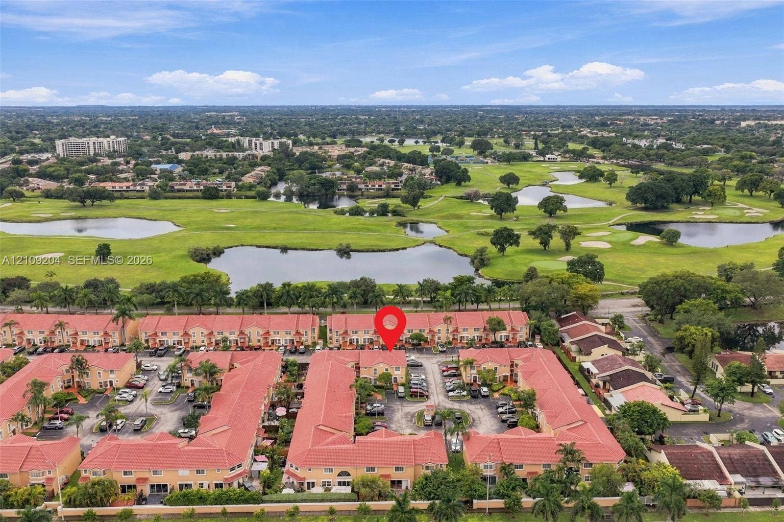 Aerial perspective of a multi-unit residential community featuring red-tile rooflines, stucco facades, and paved parking areas, situated in proximity to golf course greens and ponds