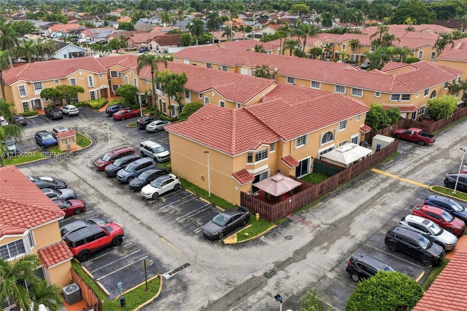 Two-story multi-family exterior featuring a terracotta-colored tiled roof, stucco facade, private fenced-in patio, and designated surface parking