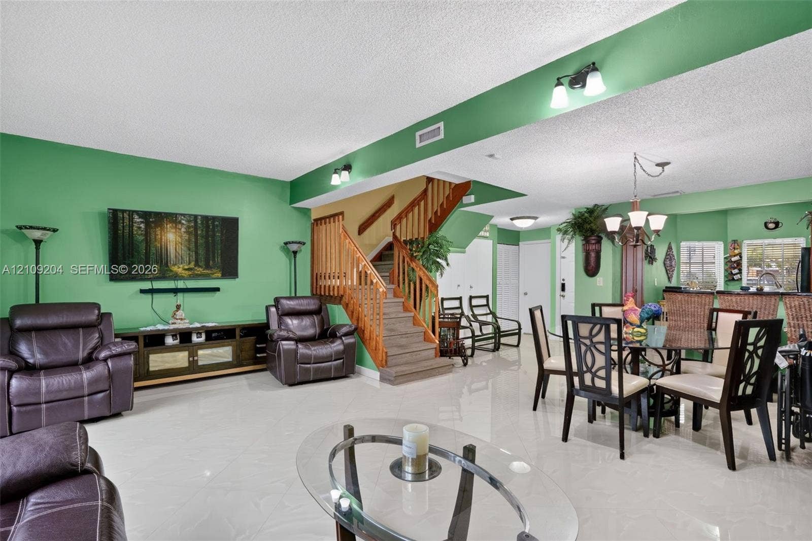 Open-concept living and dining area featuring polished light-toned tile flooring, a central staircase with wooden handrails and balusters, and multi-arm decorative chandeliers