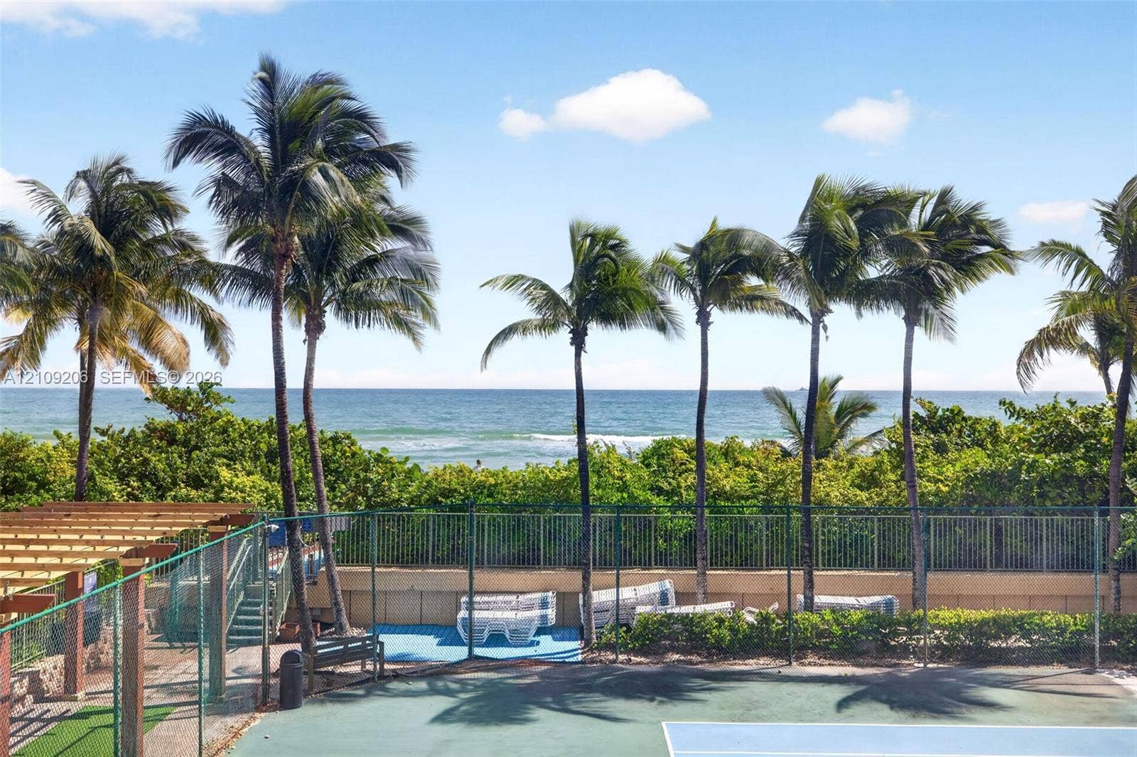 2080 S Ocean Drive, #112, Hallandale Beach, Hallandale Beach, FL 33009 Tennis court enclosed by perimeter fencing, featuring a wooden pergola, mature palm trees, and ocean frontage
