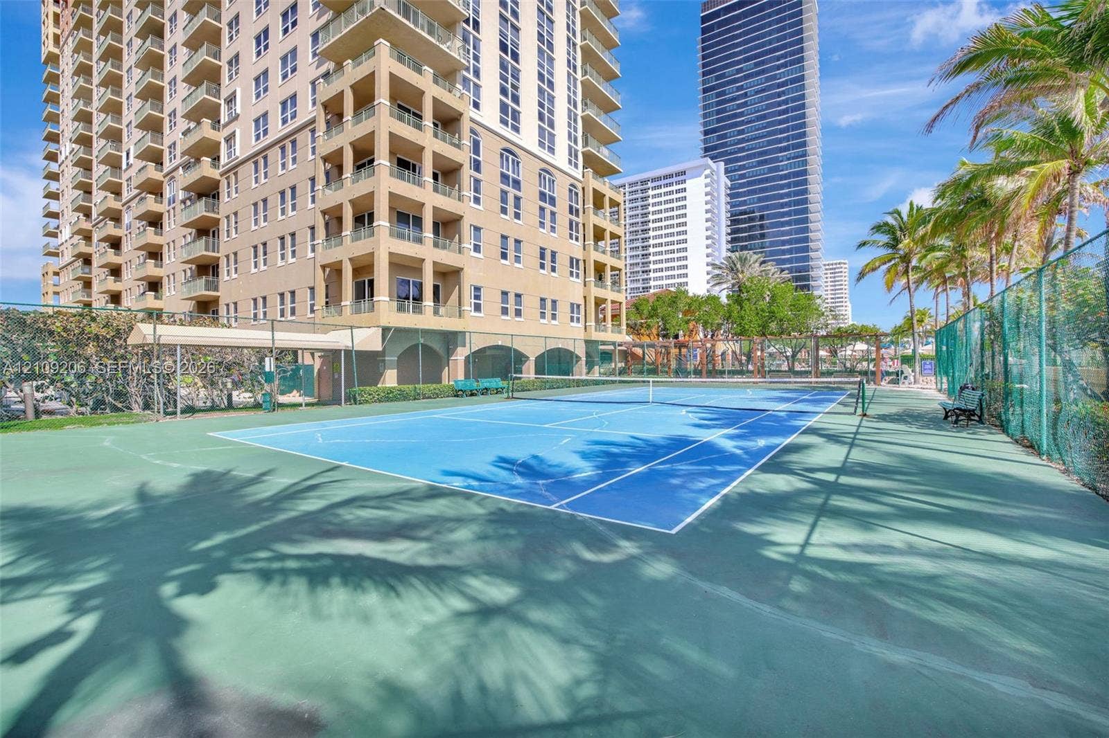 2080 S Ocean Drive, #112, Hallandale Beach, Hallandale Beach, FL 33009 Tennis court featuring a blue hard surface, chain-link fencing, and stadium-style lighting