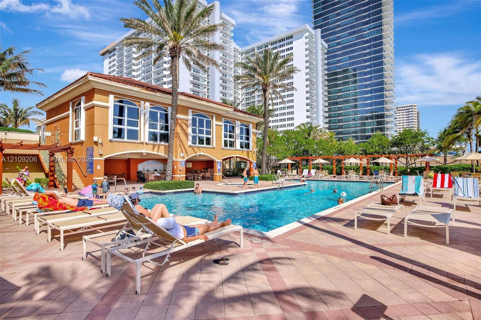 2080 S Ocean Drive, #112, Hallandale Beach, Hallandale Beach, FL 33009 Swimming pool area featuring a stone-paver deck, shaded pergola structures, mature palm trees, and an exterior clubhouse building with arched windows