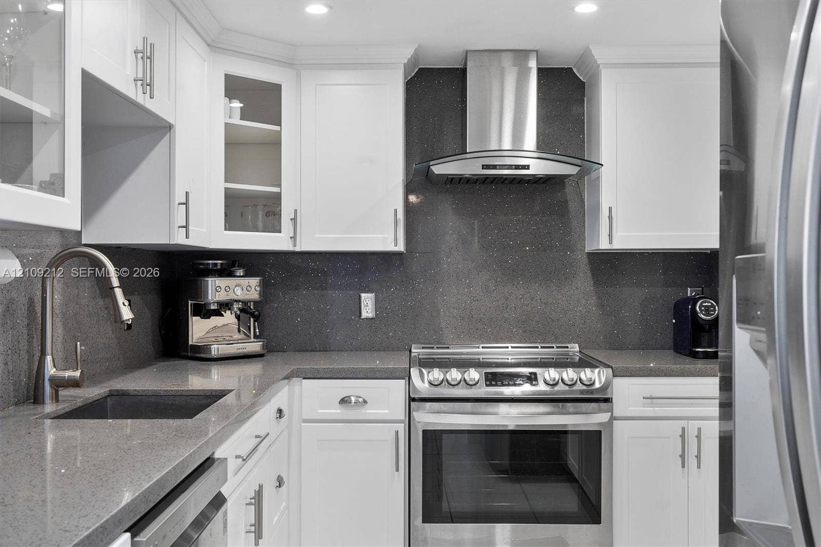 Kitchen featuring white shaker-style cabinetry, dark stone-finish countertops, full-height matching backsplash, stainless steel range with overhead chimney hood, and undermount sink with pull-down faucet