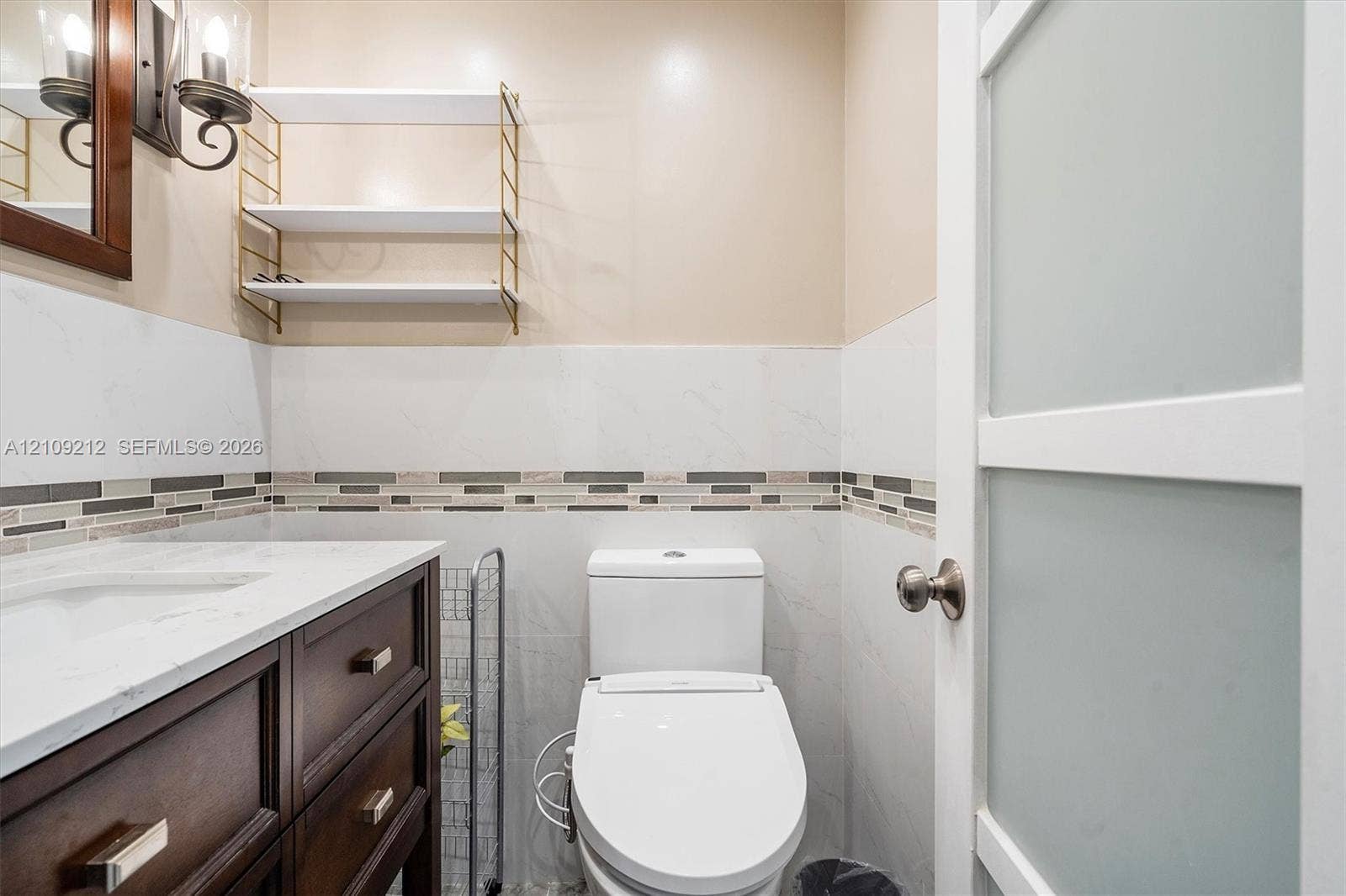 Dark wood vanity with a light-toned solid surface countertop, glass-paneled interior door, wall-mounted metal shelving, and tile wainscoting with decorative mosaic accent inlay
