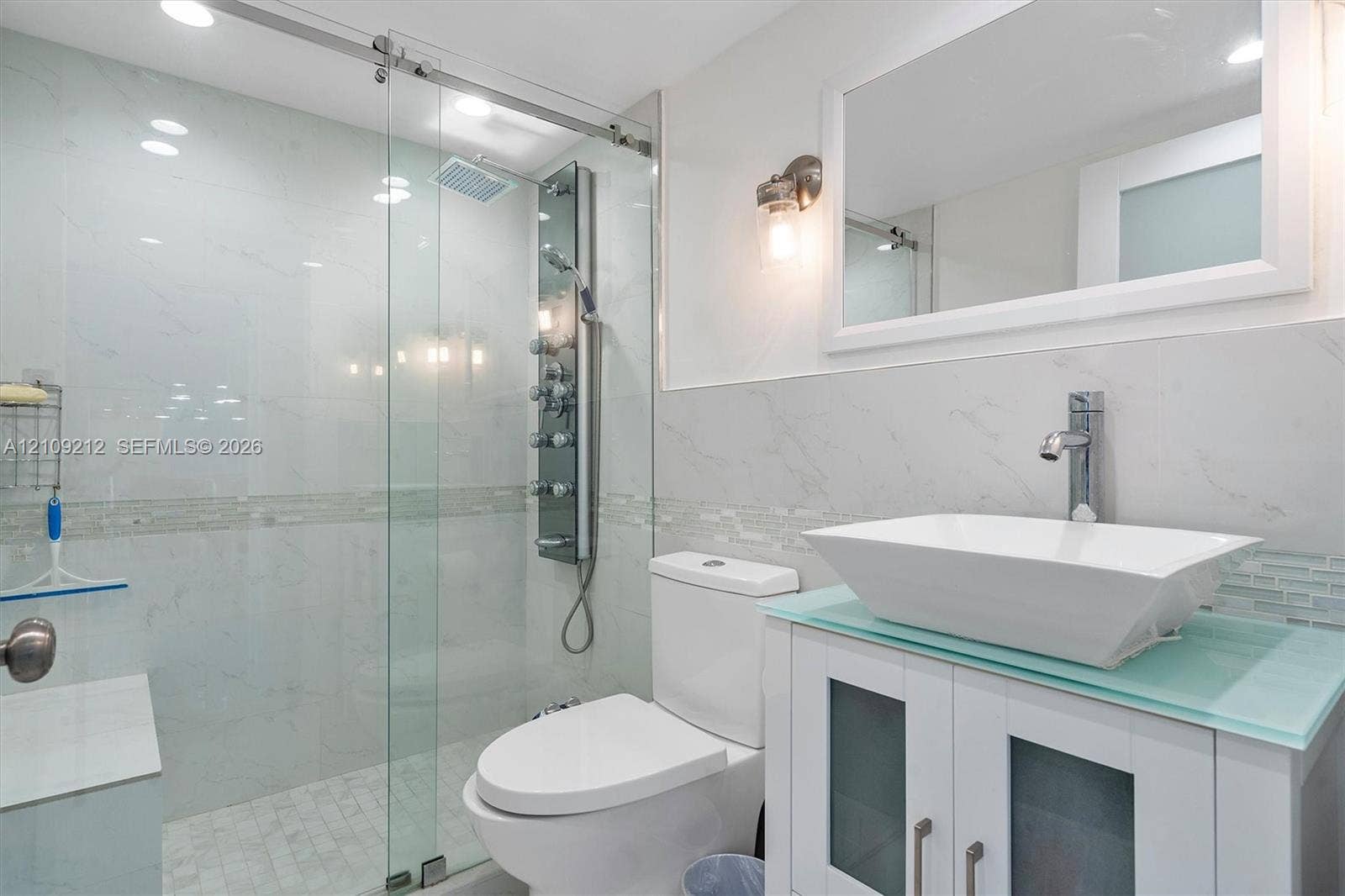 Bathroom featuring a sliding glass shower door, a multi-jet shower panel, a vessel sink with a glass countertop, and a vanity with frosted glass cabinet inserts