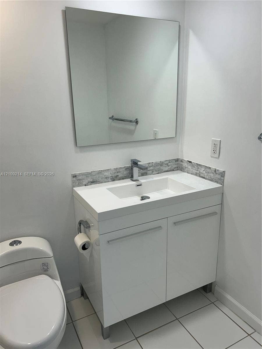 Modern vanity featuring a white finish, stone-composite countertop, integrated sink, and polished chrome hardware, complemented by a stone-tile backsplash and a frameless mirror