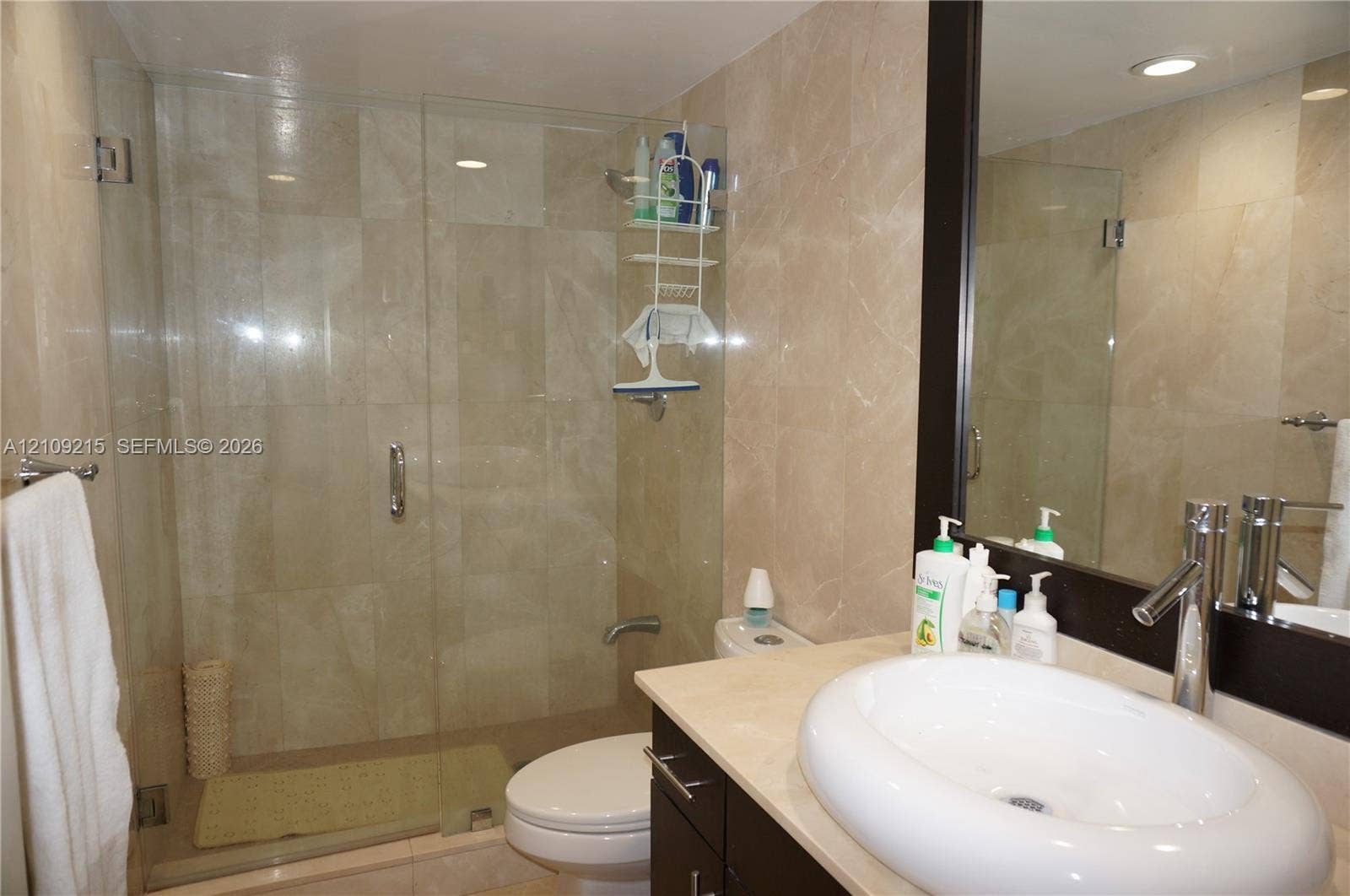 Full-wall stone tiling, glass-enclosed shower, dark-finish cabinetry, countertop-mounted oval sink, and framed mirror