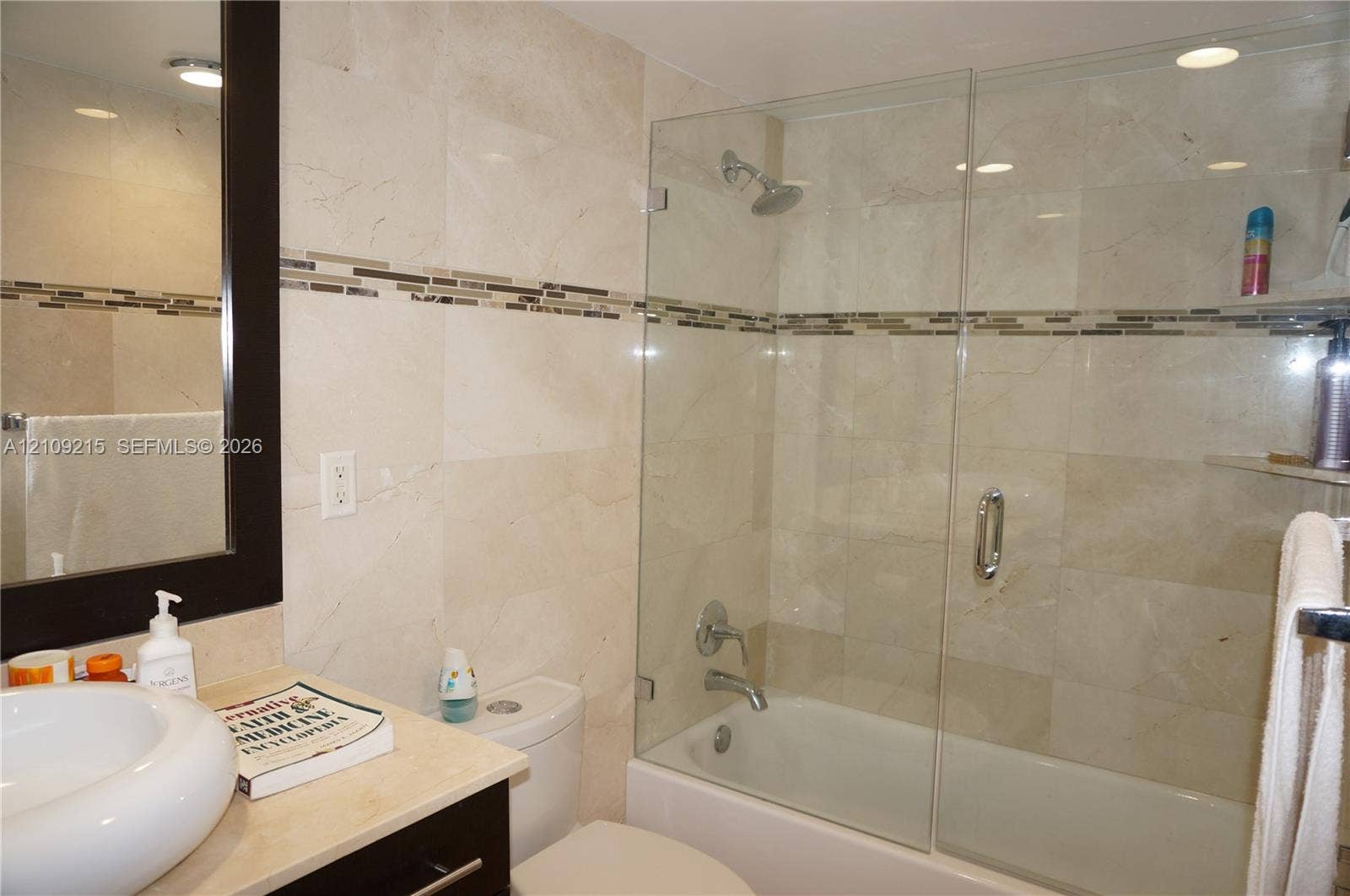 Bathroom featuring full-height stone wall tiling with decorative glass mosaic inlay, a glass-enclosed shower and tub combination, a circular vessel sink, and an integrated dark-finish vanity