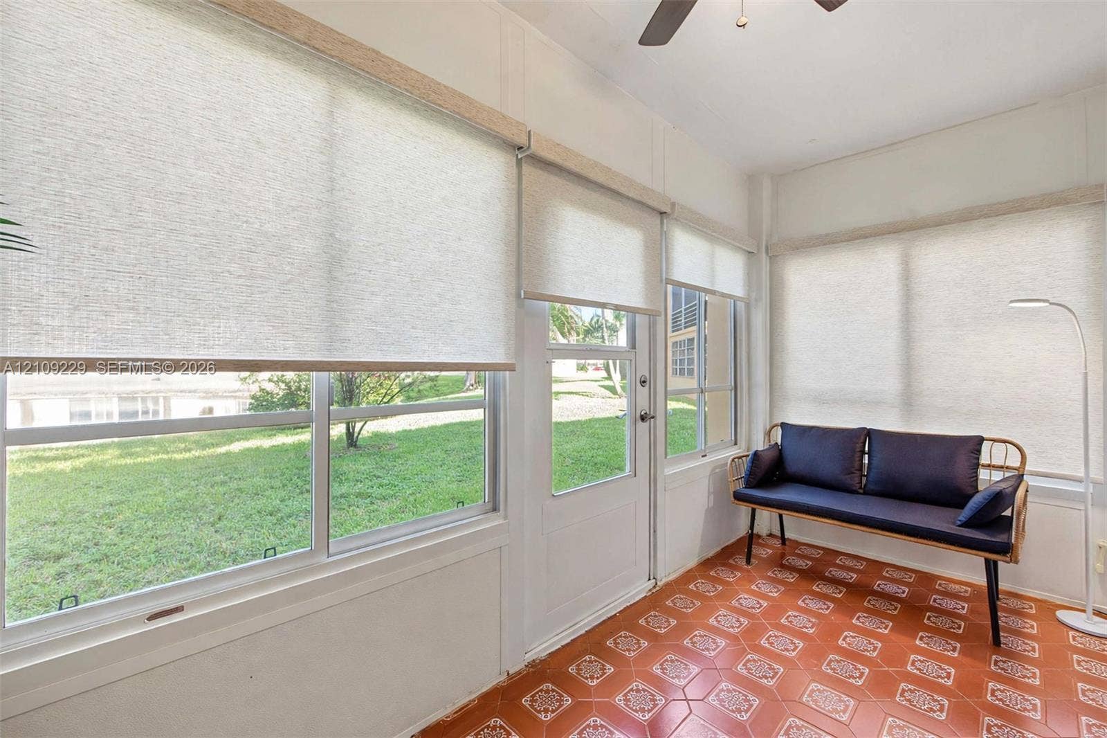 Enclosed sunroom featuring expansive windows with roller shades, patterned terra cotta tile flooring, a glass-panel exterior door, and an overhead ceiling fan