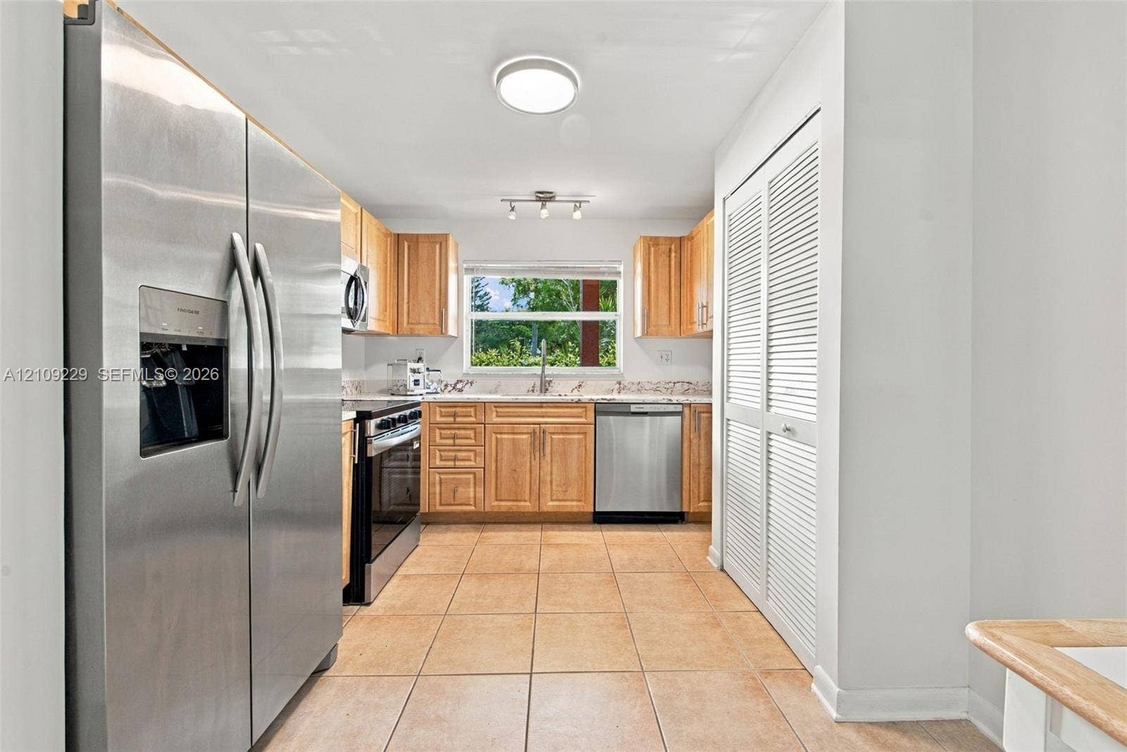 Kitchen featuring light-finish wood cabinetry, stone countertops, stainless steel appliances, ceramic tile flooring, and louvered double-door storage