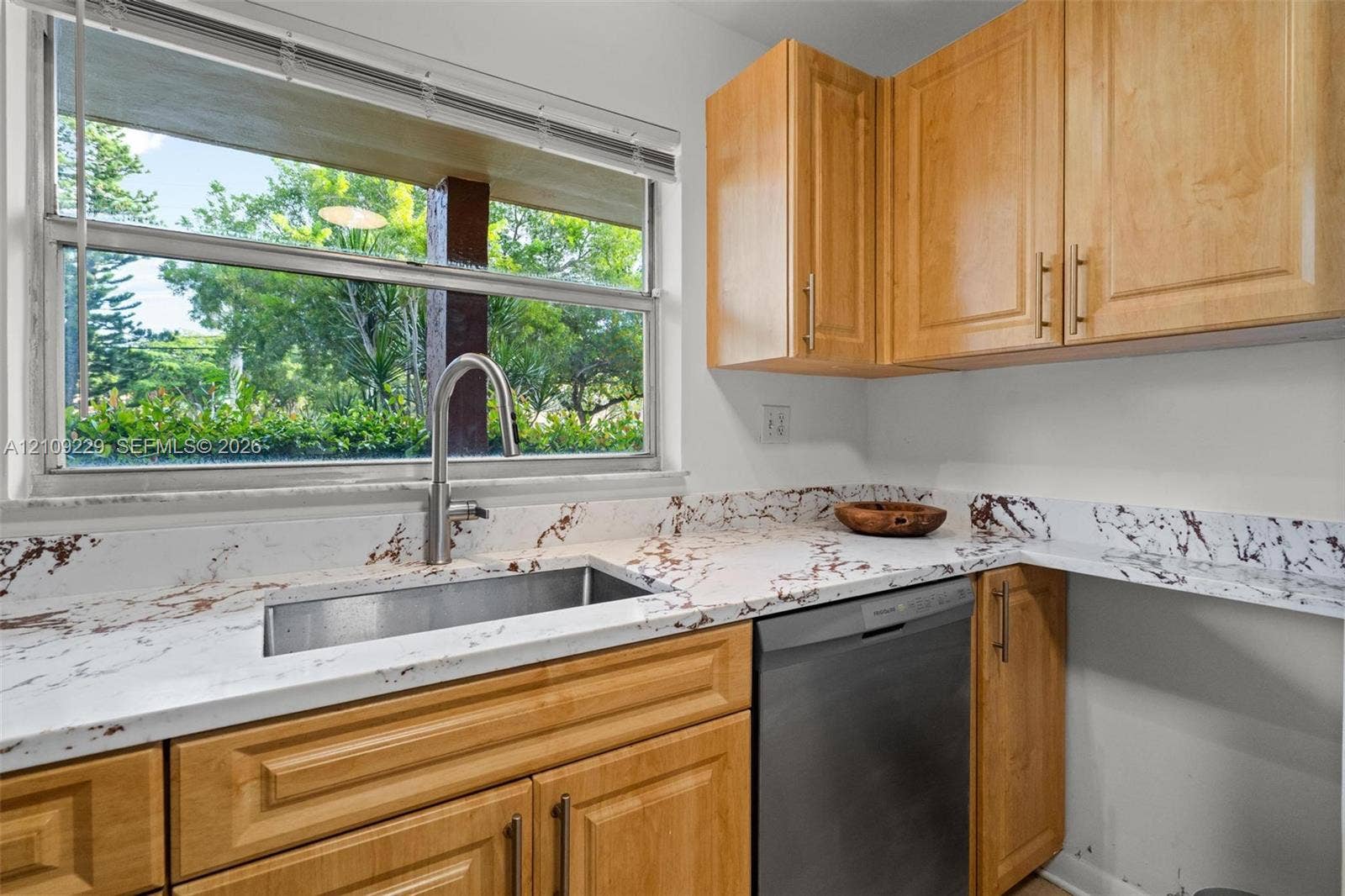 Kitchen featuring natural wood-finish cabinetry, patterned stone-surface countertops, an undermount stainless steel sink with a high-arch faucet, and an integrated stainless steel dishwasher