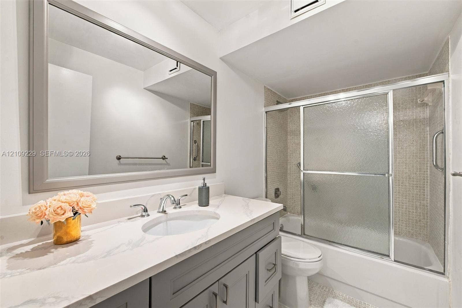 Bathroom featuring a vanity with a light-toned stone countertop, undermount sink, and brushed metal faucet, paired with a large framed mirror and a bathtub-shower combination with sliding glass doors