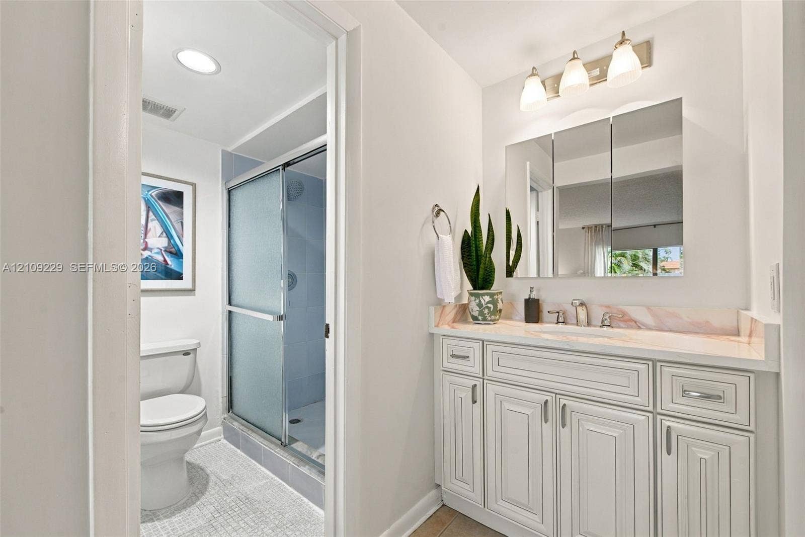 White cabinetry with recessed-panel doors, integrated sink, stone-surface vanity top, three-light wall-mounted fixture, and glass-enclosed shower