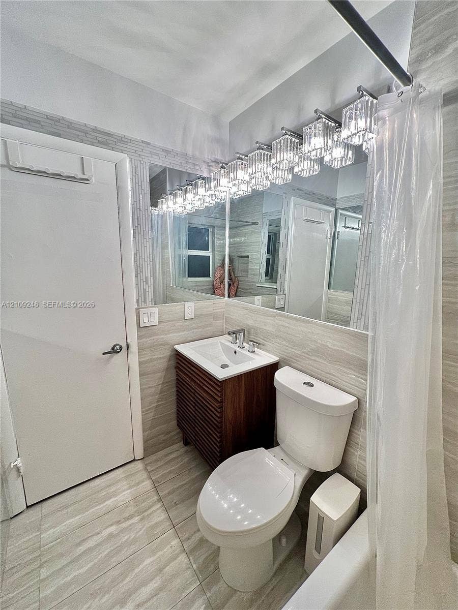 Bathroom featuring floor-to-ceiling neutral tile, wood-finish slatted vanity, integrated rectangular sink with chrome faucet, and multi-light fixture with glass prism shades