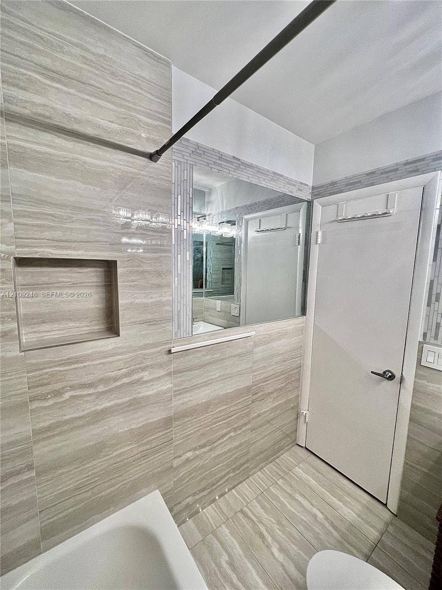 Bathroom featuring neutral-toned stone-finish wall and floor tile, recessed wall niche, glass-fronted medicine cabinet, and decorative linear mosaic tile trim
