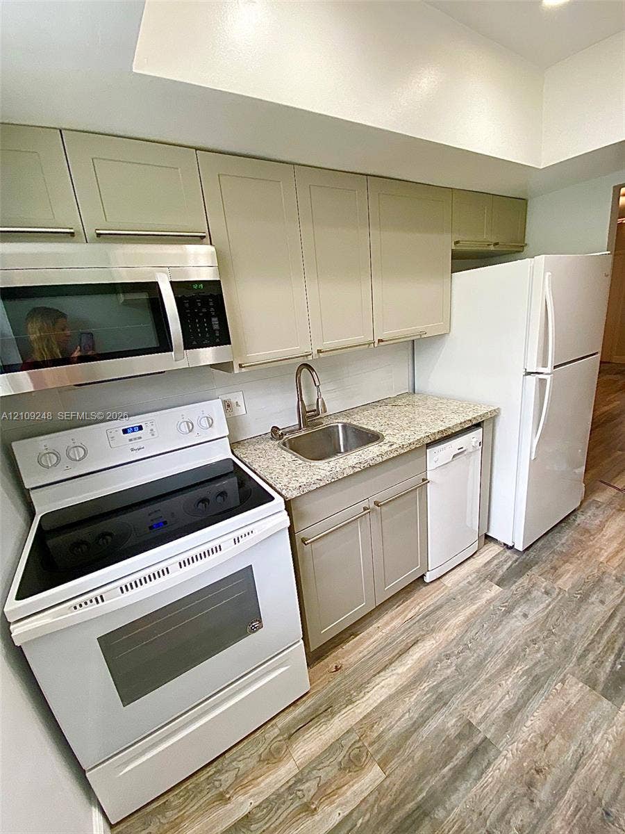 Kitchen featuring neutral-toned shaker cabinetry, stone-finish countertops, stainless steel undermount sink with gooseneck faucet, full suite of white appliances, and wood-finish flooring