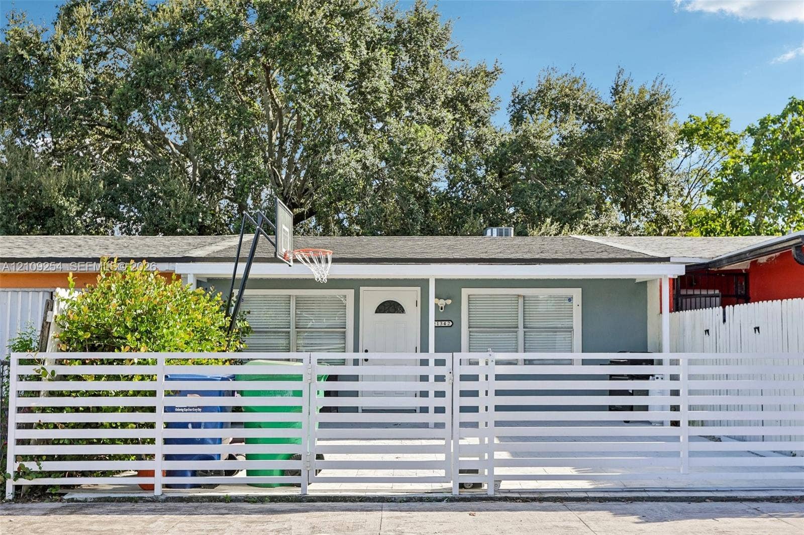 21342 NW 40th Cir Court, Miami Gardens, FL 33055 Single-story residence featuring a gray exterior, a covered front entryway, horizontal white slat fencing, and mature trees