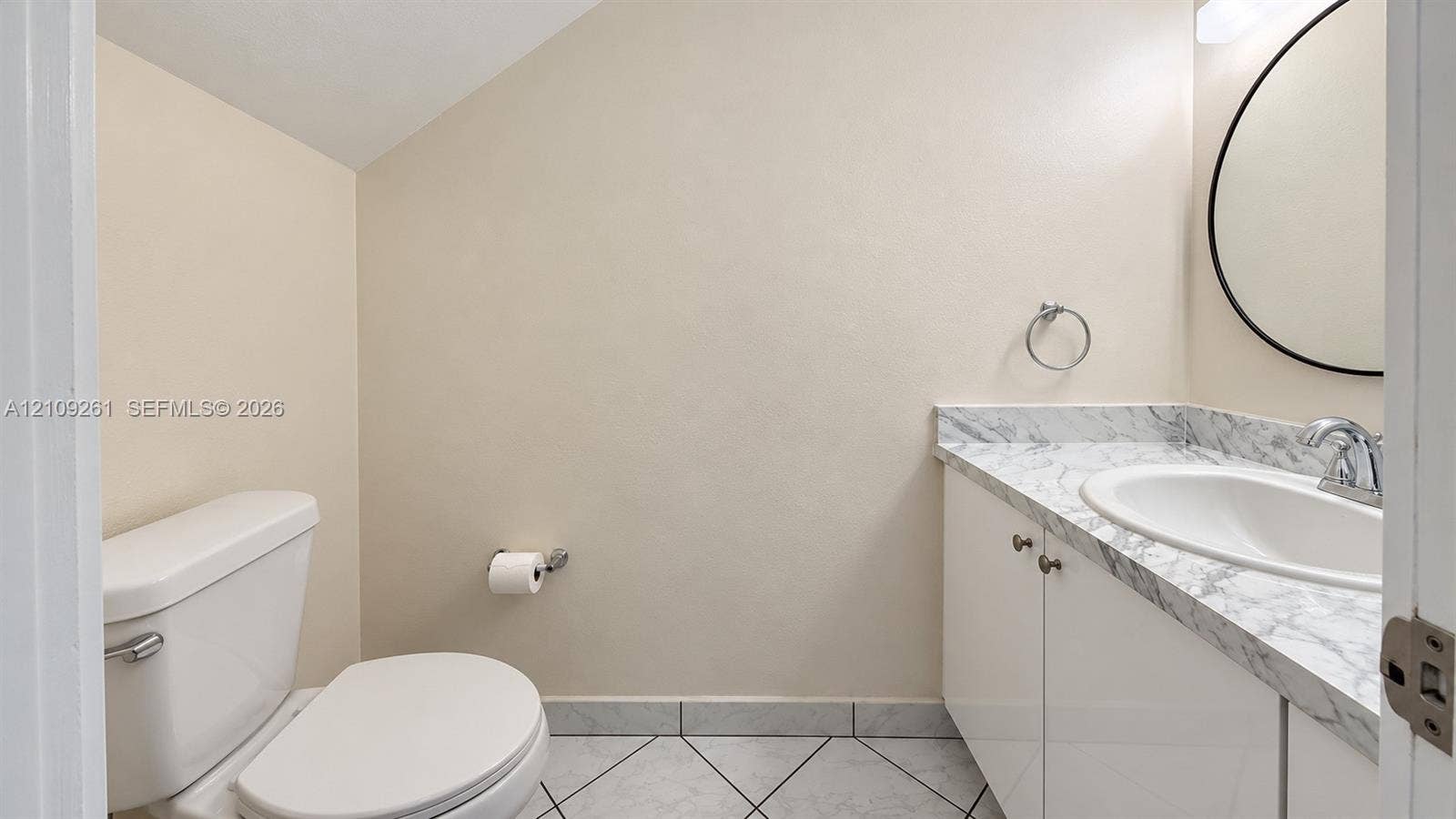 Powder room featuring a vanity with stone-finish countertop and oval basin, circular wall-mounted mirror, white cabinetry, and ceramic tile flooring