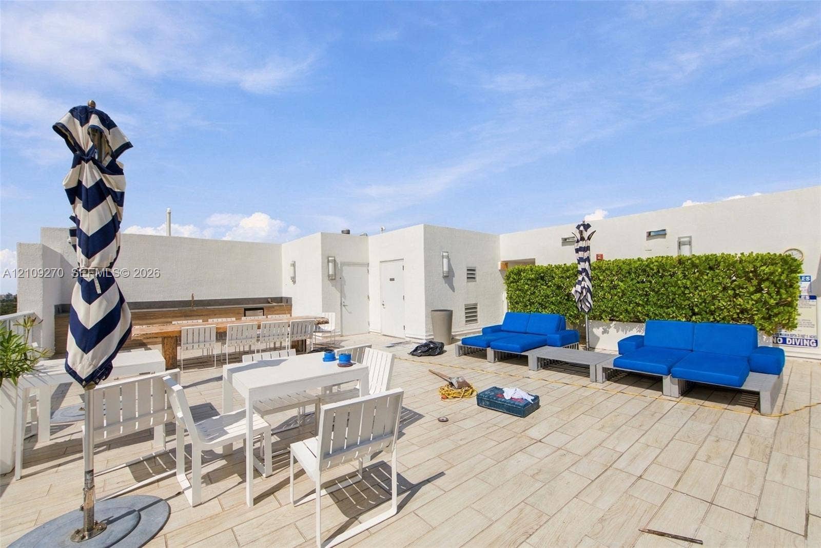 Rooftop terrace featuring light-toned tile flooring, integrated perimeter hedge, stucco wall finishes, and built-in outdoor kitchen structure with attached wall-mounted lighting