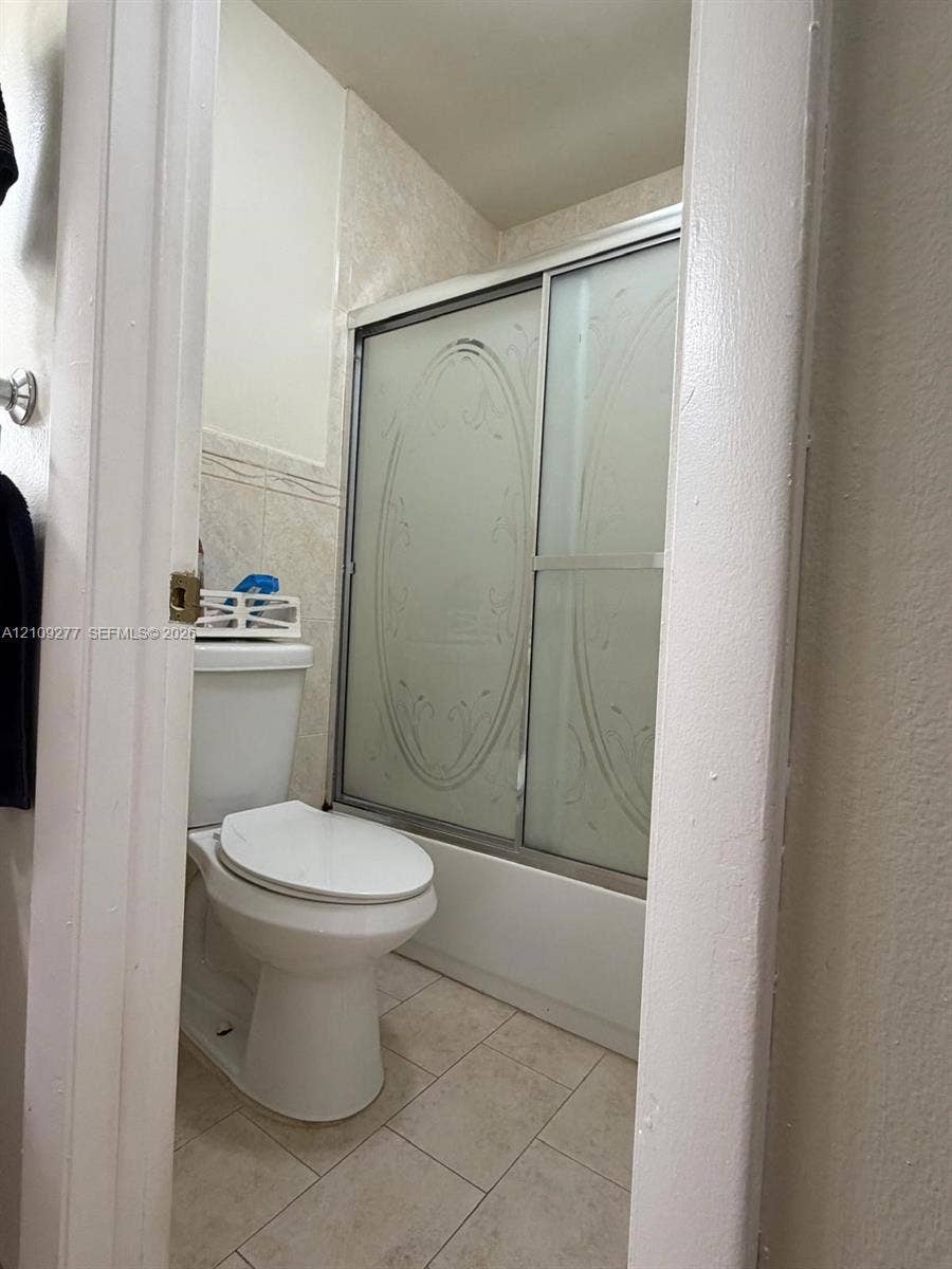 Full bathroom featuring a shower-tub combination with sliding etched glass doors, tile surround, porcelain toilet, and neutral tile flooring