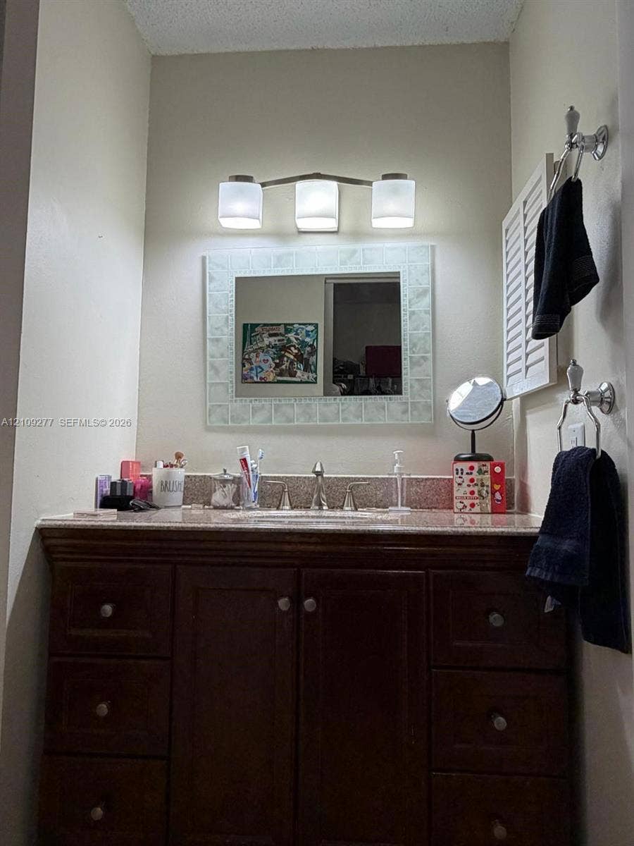 Vanity featuring dark wood cabinetry, integrated stone-finish countertop with an undermount basin, three-light fixture with frosted glass shades, and a decorative tile-framed mirror
