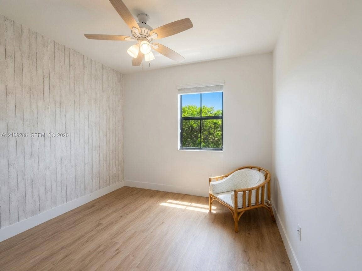 Room featuring wood-finish flooring, a wood-paneled accent wall, a multi-blade ceiling fan with integrated lighting, and a single-pane window